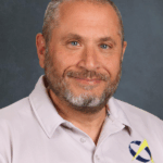 A man with a beard smiles, wearing a light gray polo shirt with a logo on the left side. Background is dark.