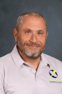 A man with a beard smiles, wearing a light gray polo shirt with a logo on the left side. Background is dark.