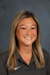 A smiling woman with long, straight hair wearing a dark polo shirt poses against a textured gray background.