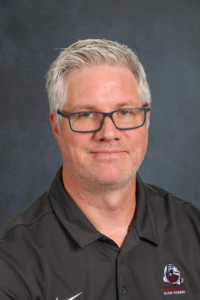 Portrait of a man with short gray hair and glasses, wearing a dark shirt with a school logo, smiling against a gray backdrop.