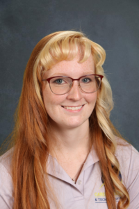 A smiling woman with long, red hair and glasses, wearing a light gray shirt with a logo.