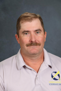 A man with a mustache wearing a light gray polo shirt, standing against a dark background.