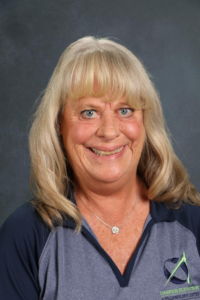 Smiling woman with long, wavy blonde hair, wearing a dark polo shirt with a logo, against a gray background.