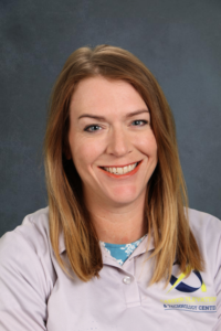 A woman with long, light brown hair smiles, wearing a gray polo shirt with a logo on the chest. Background is gray.